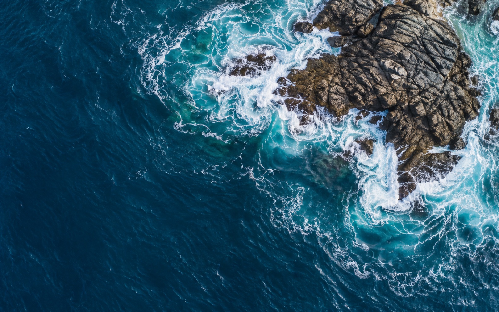An overhead study of deep blue water and white sea foam breaking around a rocky outcrop.
